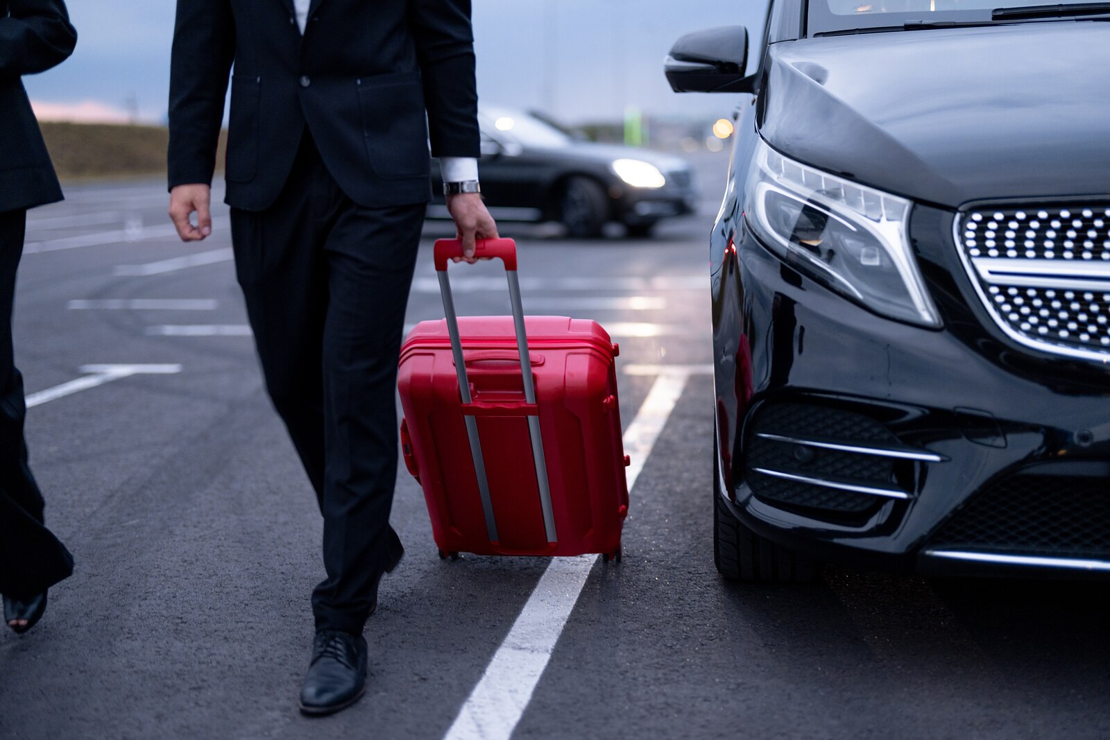Chauffeur in a dark suit pulling a red suitcase past a black Mercedes van in a parking lot