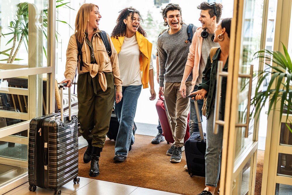Group of young friends laughing as they arrive at a hotel entrance with rolling suitcases